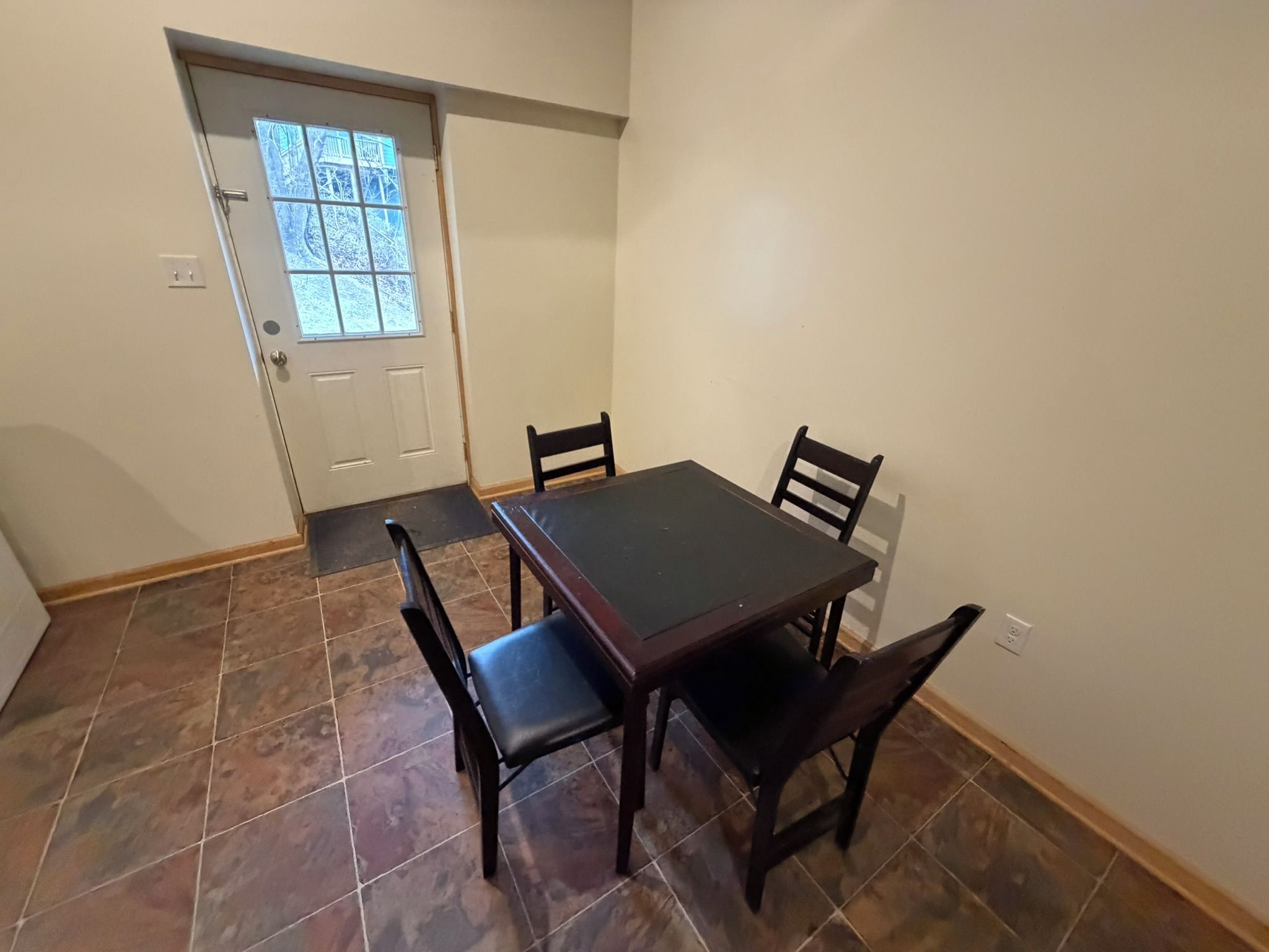 Un comedor con una mesa de madera oscura y cuatro sillas, una puerta con ventana, y piso de baldosas.
