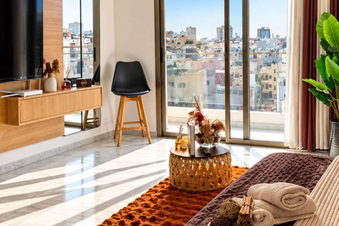 A modern living room with a large TV, a black chair, a round coffee table with a vase, and a view of a cityscape through large windows.