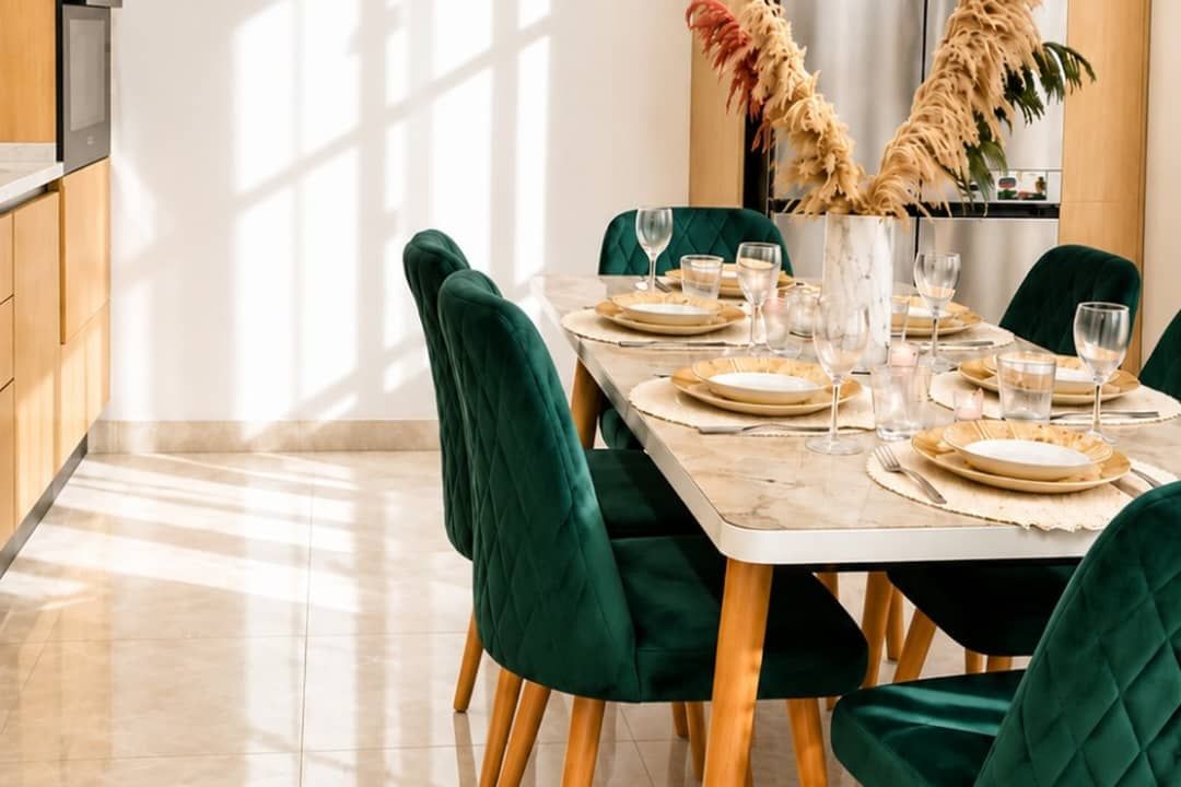 Un comedor con una mesa dispuesta para cuatro, sillas tapizadas en verde y un florero con plantas secas.