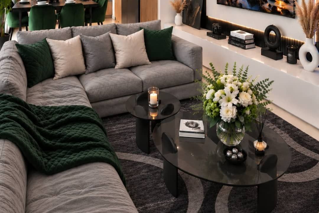 A modern living room with a grey sectional sofa, a round coffee table with a candle, and a vase with white flowers.