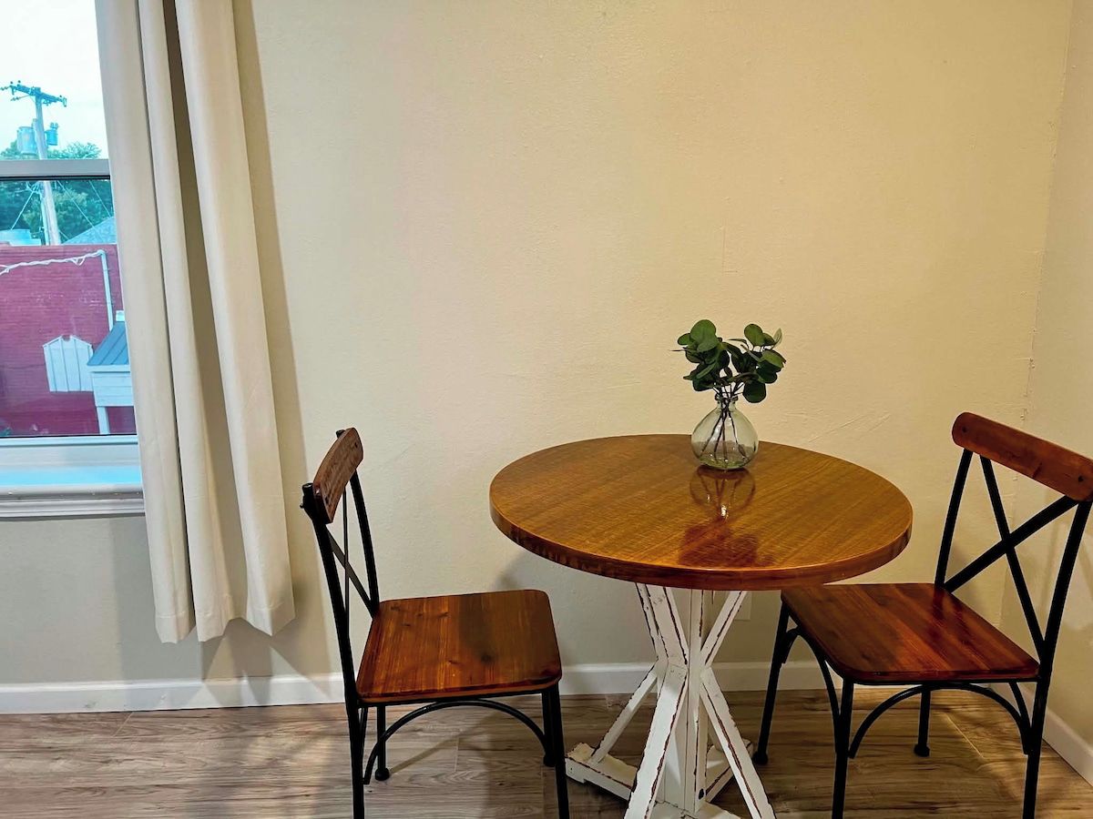 Une table ronde en bois, accompagnée de deux chaises, un vase avec une plante et une fenêtre avec des rideaux.