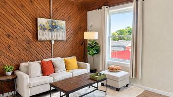 A living room with a wooden ceiling, a sofa, a coffee table, a floor lamp, a painting, and a window with curtains.