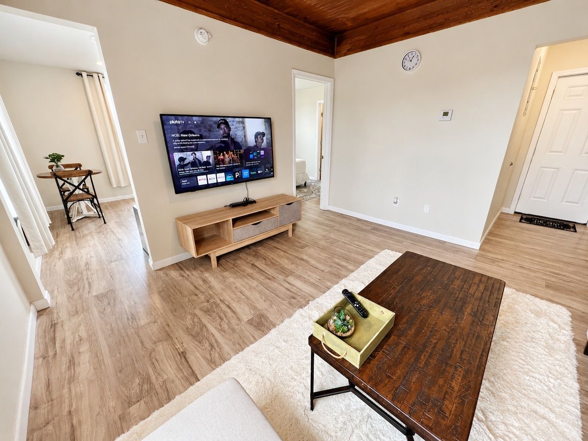Un soggiorno con pavimento in legno, una TV fissata al muro, un tavolino in legno e un tappeto bianco.