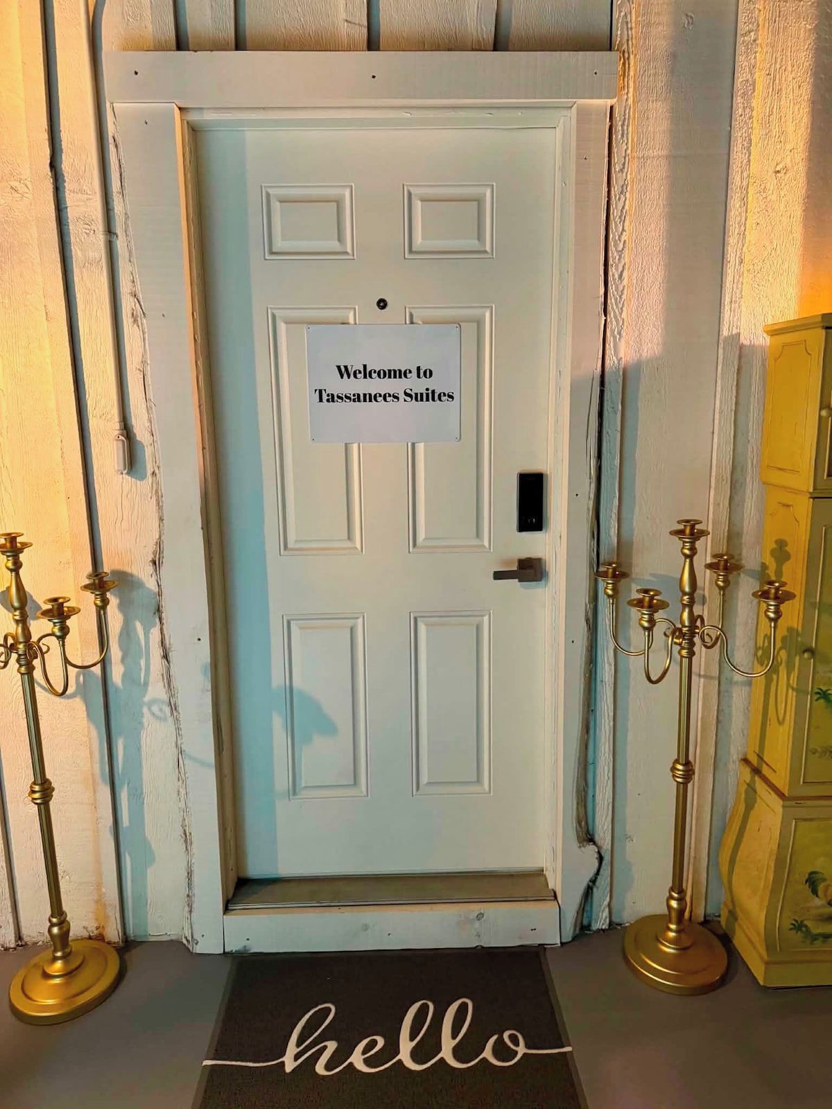 Eine weiße Tür mit einem Schild, auf dem 'Welcome to Tassanees Suites' steht, und einer Fußmatte mit dem Wort 'hello'.