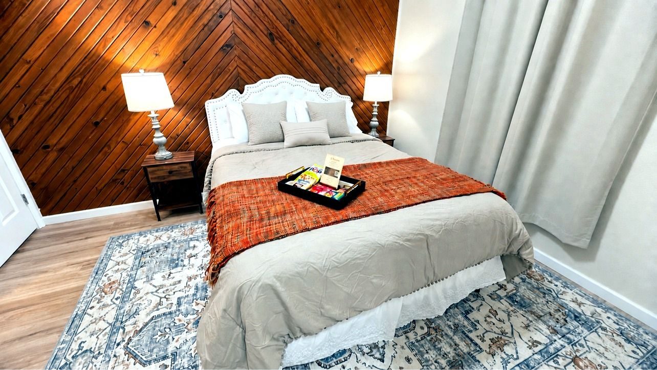 A hotel room with a wooden ceiling, a bed with a beige bedspread, two bedside lamps, a tray with magazines, and a patterned rug.