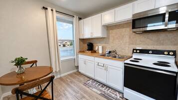 A compact kitchen with white cabinets, a stainless steel microwave, a black stove, and a wooden dining table with chairs.