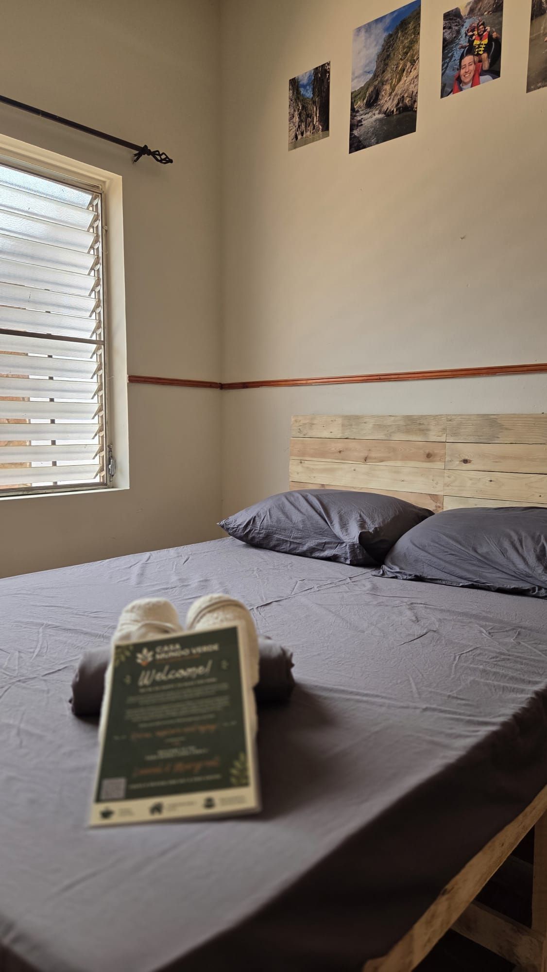 Una camera da letto con una testiera in legno, un letto con biancheria grigia, una finestra con tende e un cartello di benvenuto sul letto.