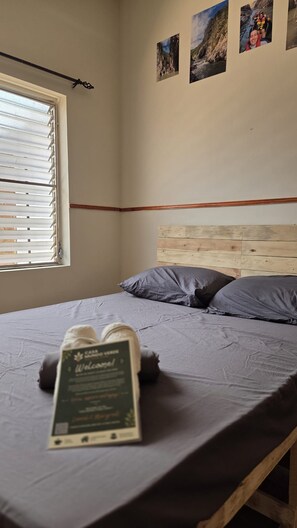 A bedroom with a wooden headboard, a bed with grey bedding, a window with blinds, and a welcome sign on the bed. - Casa Mundo Verde - Local Hostel (LEÓN)