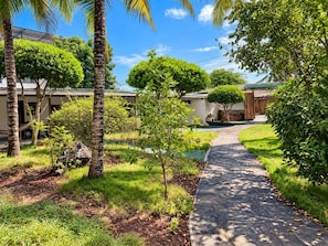A pathway leading to a building with a covered outdoor area, surrounded by greenery and palm trees.