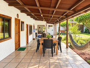 A covered patio with a dining table and chairs, a hammock, and a view of a garden.