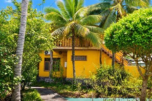 A yellow building with a wooden staircase, surrounded by lush greenery and palm trees.