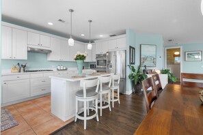 A modern kitchen with white cabinetry, a central island with bar stools, and a dining area with a wooden table and chairs. - Skyline Serenity- Heated Pool & Water Views - 3 story Coastal Retreat (Jensen Beach)