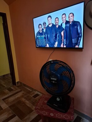 A television mounted on a wall displaying a group of people. Below the TV, there is a black fan on a small table. - New Listing (Vera Cruz)