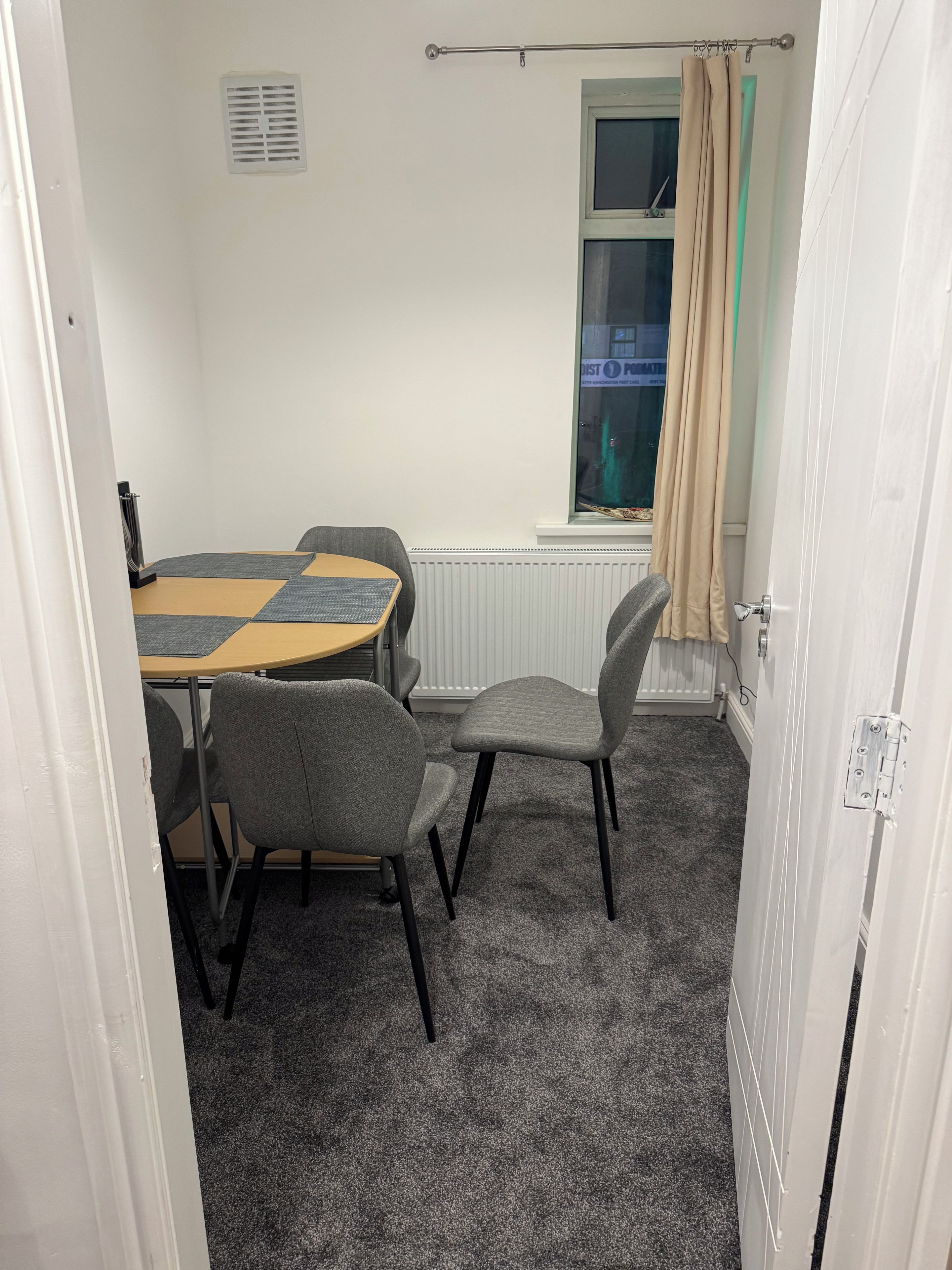 A room with a round table and four chairs, a radiator, a window with a curtain, and a door.
