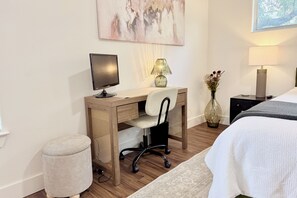 Habitació d'hotel amb escriptori de fusta, un monitor d'ordinador, una cadira, una taula de nit amb una làmpada, un vas amb flors i un llit amb mantell blanc.