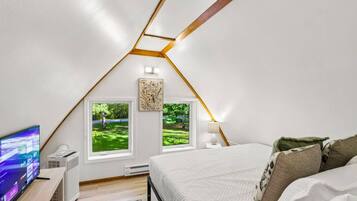 Un dormitorio en el ático con techo inclinado, dos ventanas con vistas a la vegetación, una cama con almohadas, un televisor sobre un soporte y un calentador.