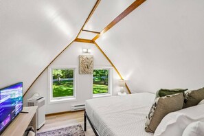 Een zolderkamer met schuine wanden, twee ramen met uitzicht op groen, een bed met kussens, een televisie op een standaard en een verwarming.