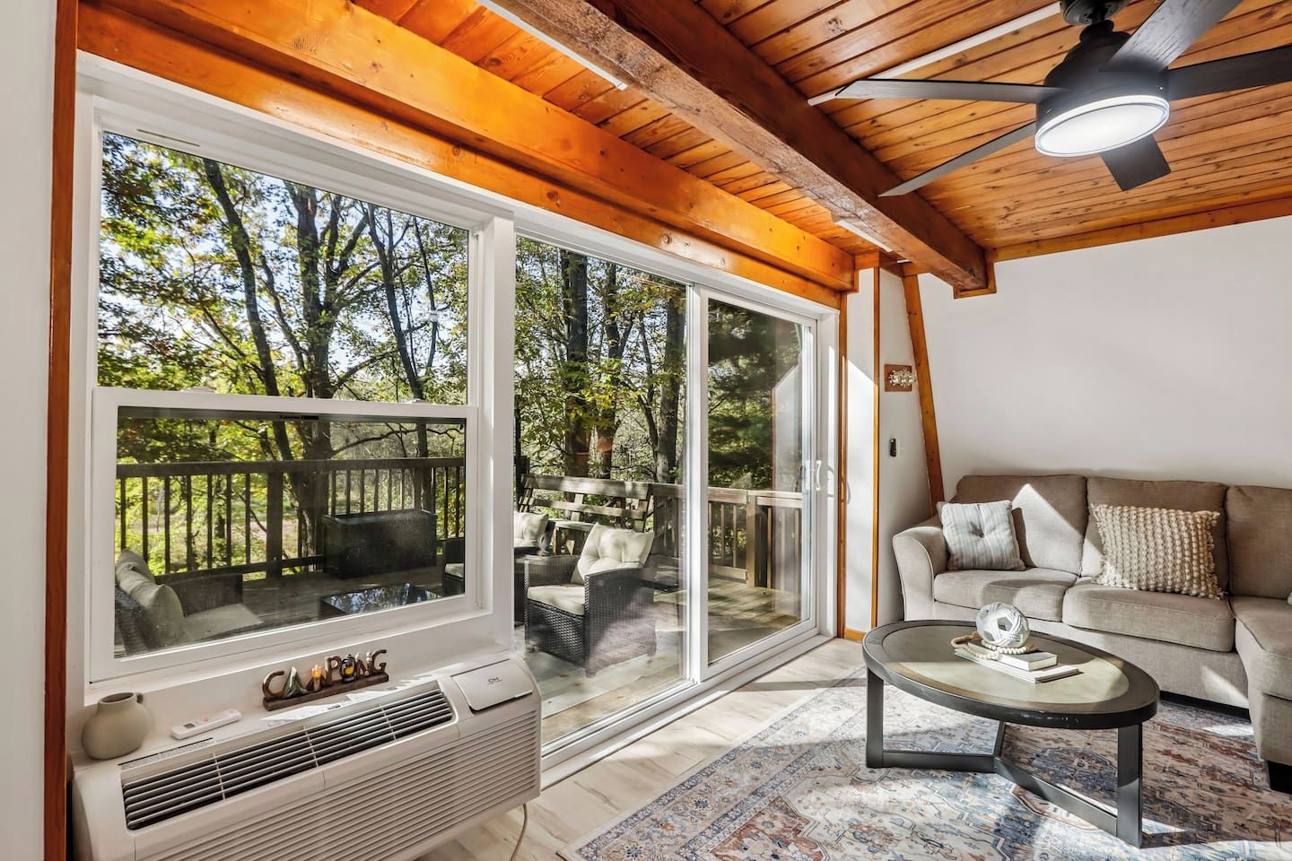 Una habitación con un amplio ventanal corredera que da a una terraza exterior, un sofá color beige, una mesita de centro y un ventilador de techo.