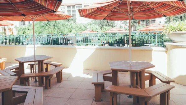 Un patio al aire libre con mesas y bancos de madera, sombrillas a rayas y vistas a zonas verdes y edificios al fondo.