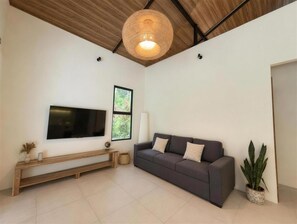 A modern living room with a flat-screen TV mounted on the wall, a grey sofa with cushions, a wooden bench, a large woven light fixture, and a potted plant. - New Listing (Cocles)