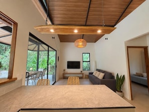 Una sala de estar moderna con una mesa grande, un sofá y un televisor. Hay ventanas amplias y un techo con vigas de madera.