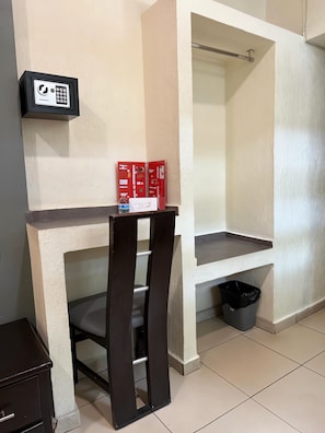 Una habitación con un escritorio, una silla, un mueble de almacenamiento rojo, un cubo de basura negro y un panel de control fijado a la pared.