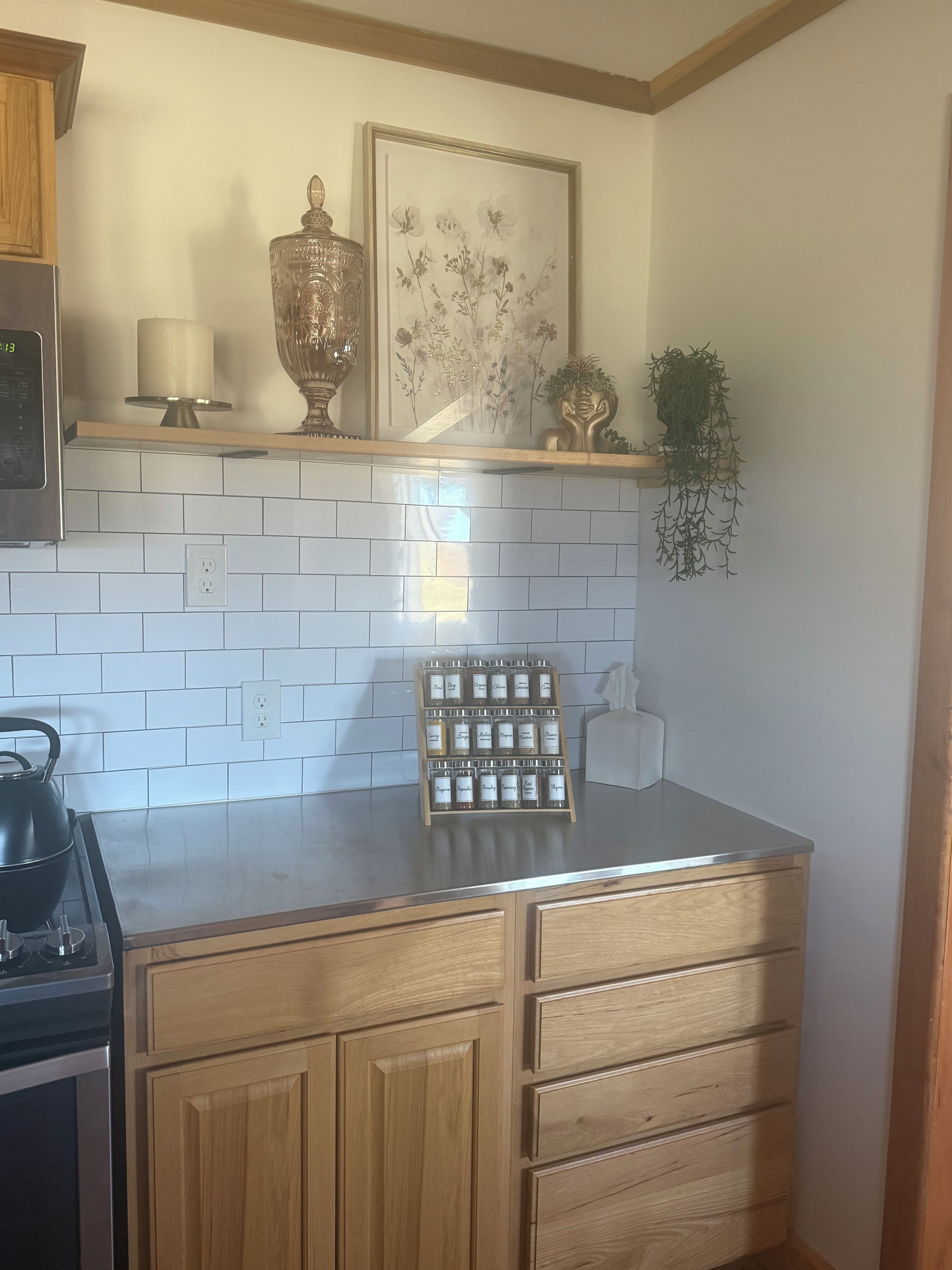 Una cocina con baldosas blancas tipo metro, estufa de acero inoxidable, armarios de madera, una obra de arte floral enmarco y una pequeña planta en maceta.