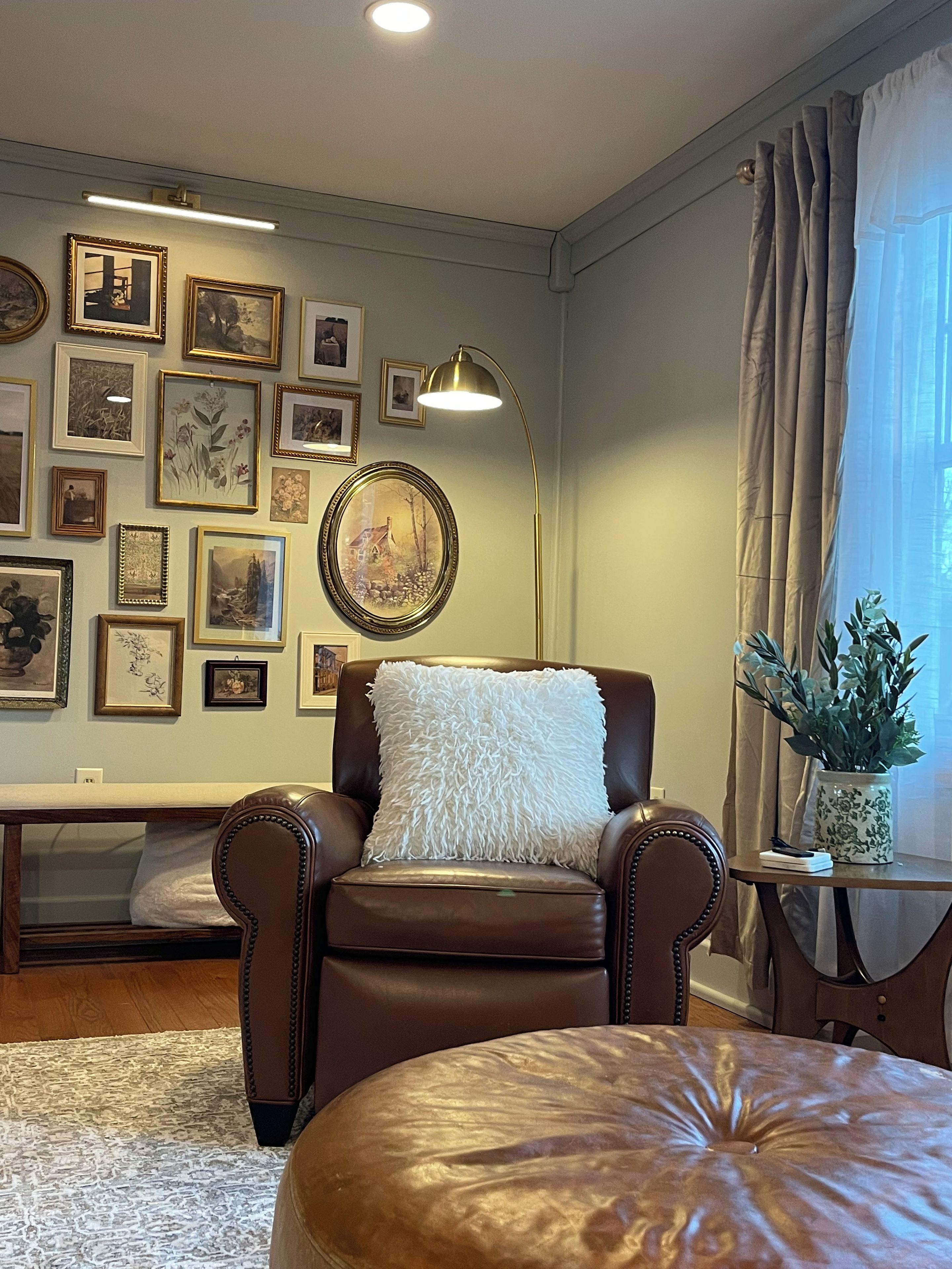 Un salón con un sillón de cuero marrón, un almohadón blanco mullido, un reposapiés redondo y una pared decorada con cuadros.