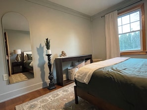Un dormitorio con cama, mesita de noche, cómoda, espejo, ventana con cortinas y una planta en maceta.