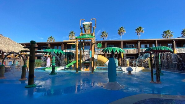 A water park with slides and a pool area, surrounded by palm trees and a hotel building.