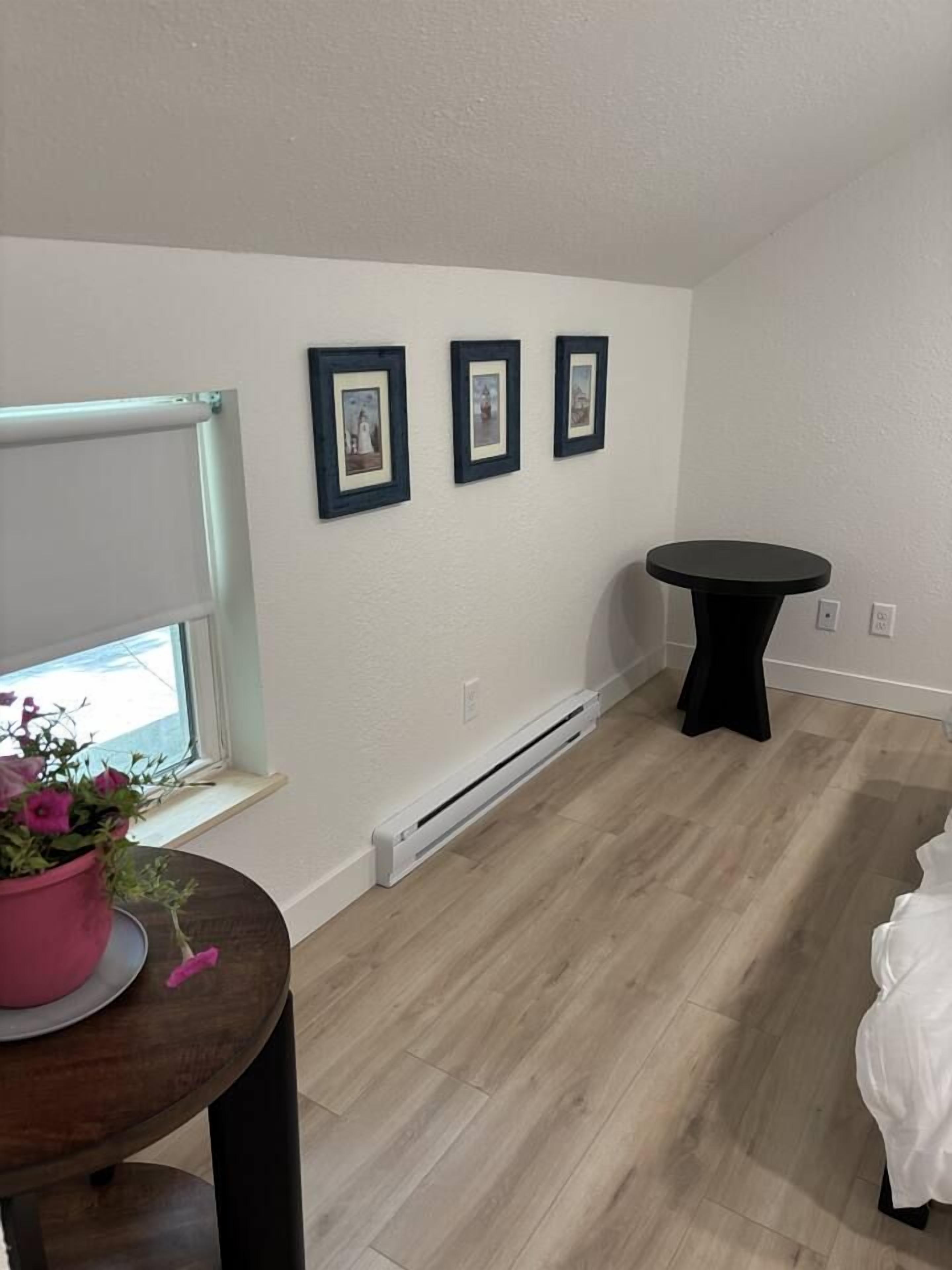 A room with a wooden floor, a window with a view, a small round table, and three framed pictures on the wall.