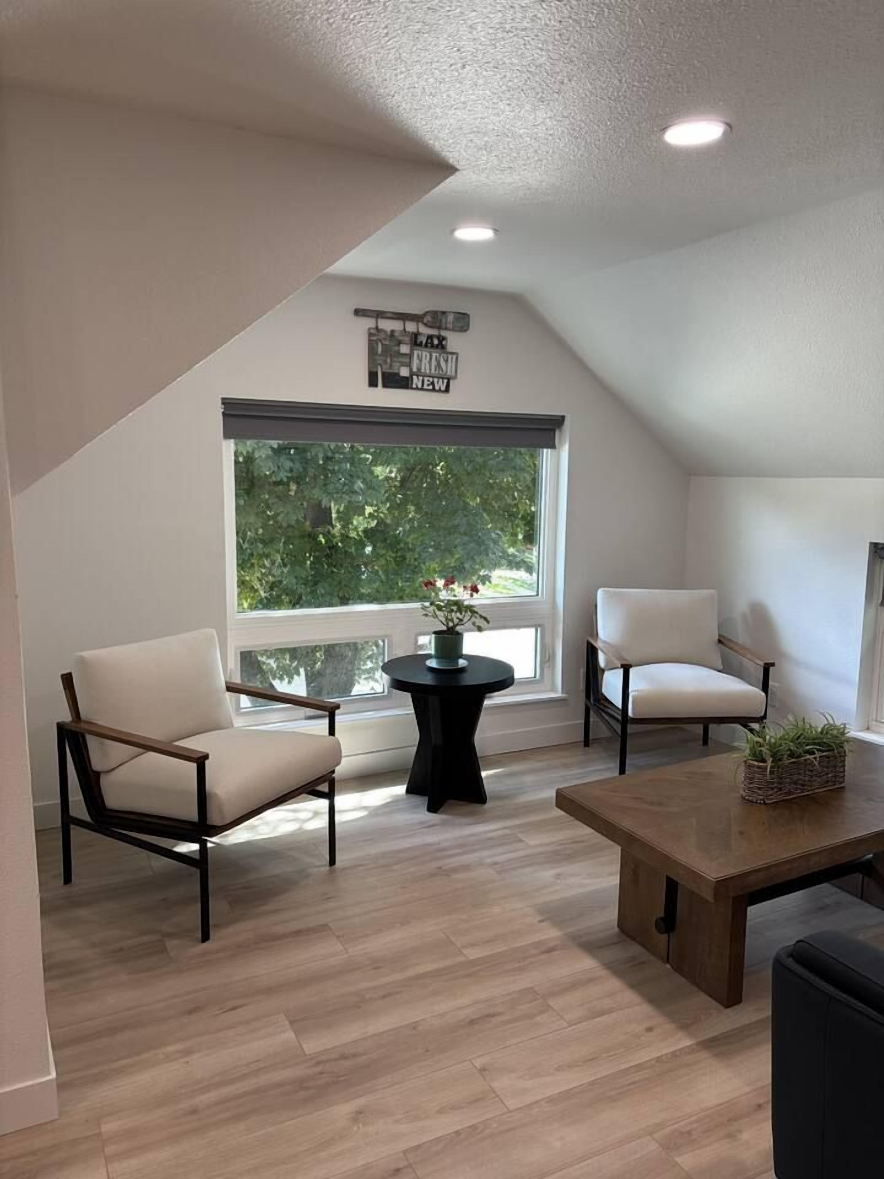 A modern loft bedroom with a sloped ceiling, a large window, two white chairs, a wooden table, and a small side table with a plant.