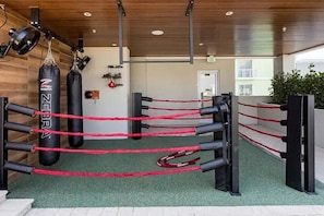 Un ring de boxeo con cuerdas y un saco de golpes, paredes de madera y una puerta al fondo.