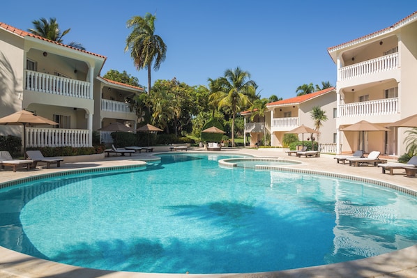 A swimming pool with lounge chairs and umbrellas, surrounded by multi-story buildings with balconies and palm trees. - 2BR Suite with Kitchen in All-Inclusive Resort, Half Block to Cofresi Beach (Puerto Plata)