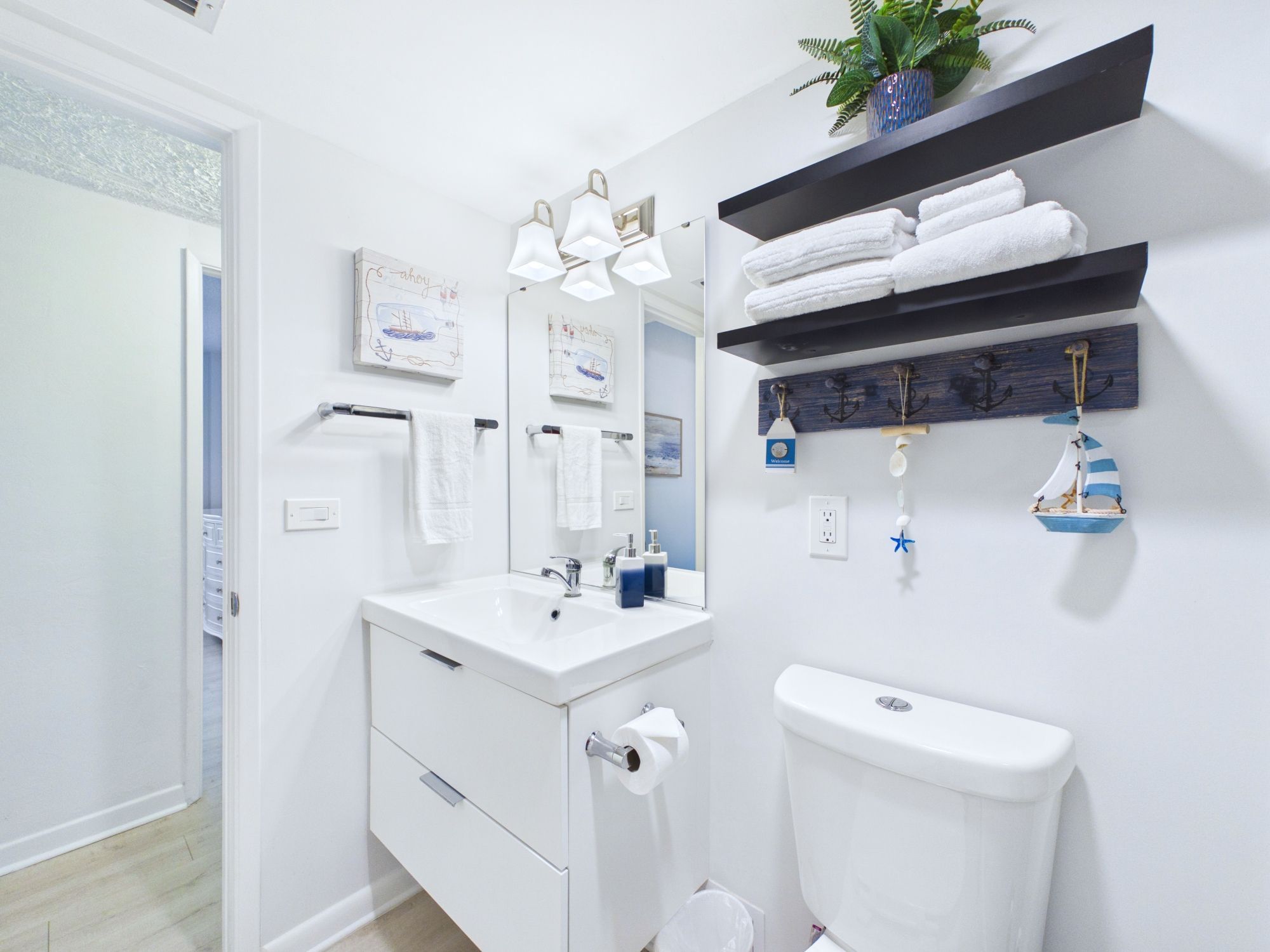 A modern bathroom with a white sink, mirror, and toilet. There are towels on a shelf, a potted plant, and a decorative anchor.