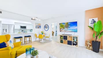Una sala moderna con un sofá amarillo, un comedor con sillas amarillas, una cocina con armarios blancos y un cuadro grande con una escena de playa.