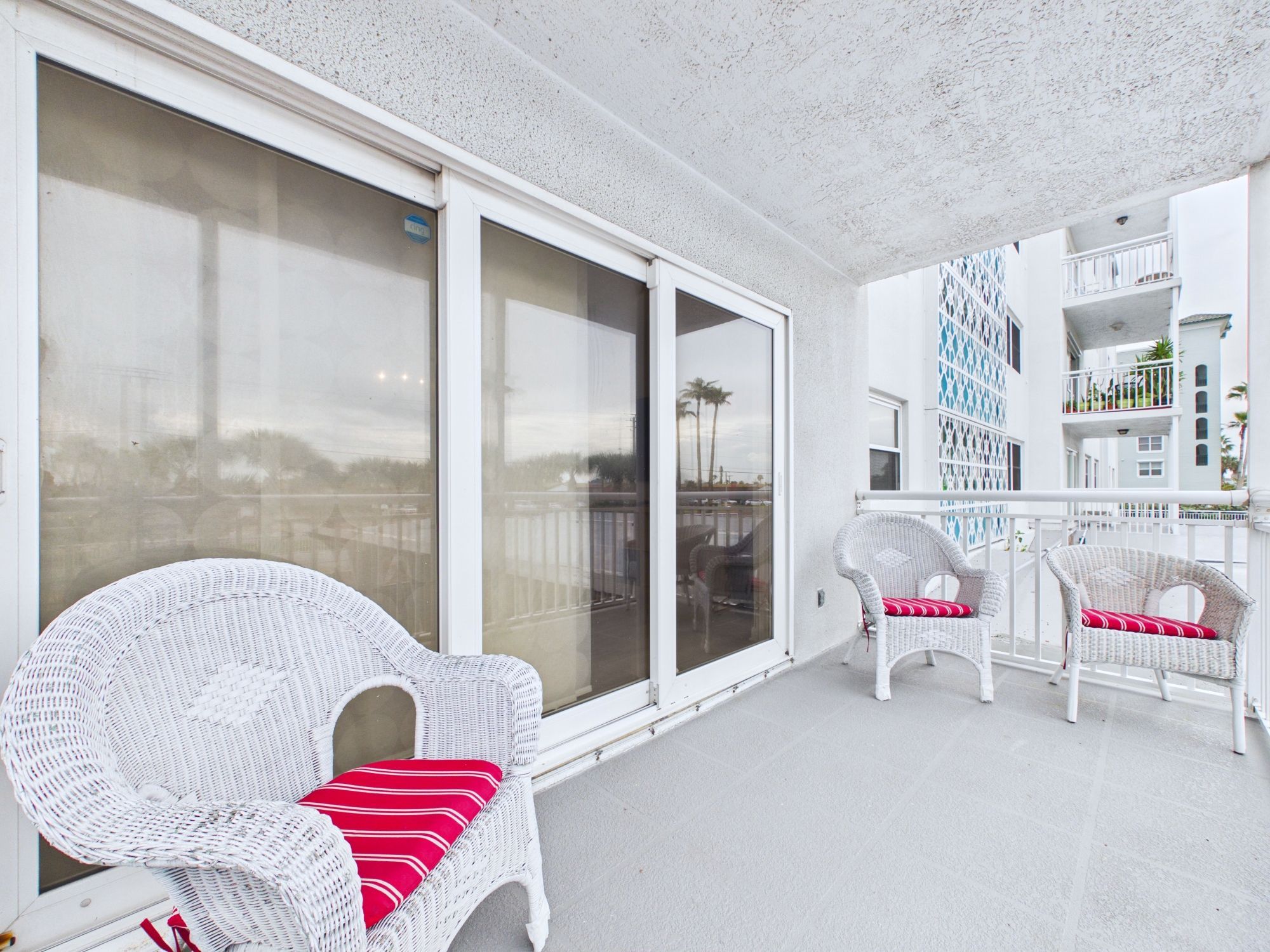Un balcón con muebles de mimbre blanco y un cojín rojo, un edificio blanco con fachada decorativa y una vista despejada del cielo.
