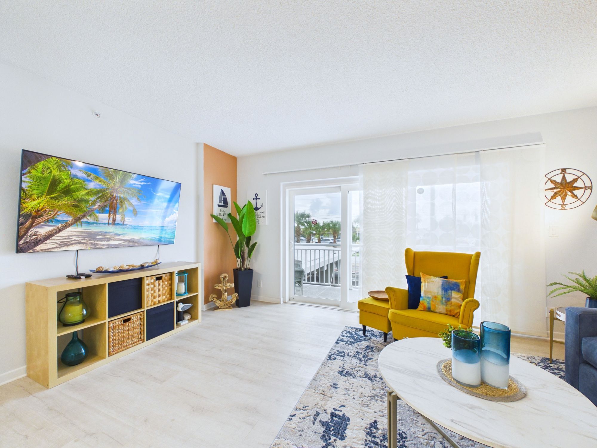 A living room with a flat-screen TV, a yellow armchair, a coffee table, and a large window with a view of the outdoors.