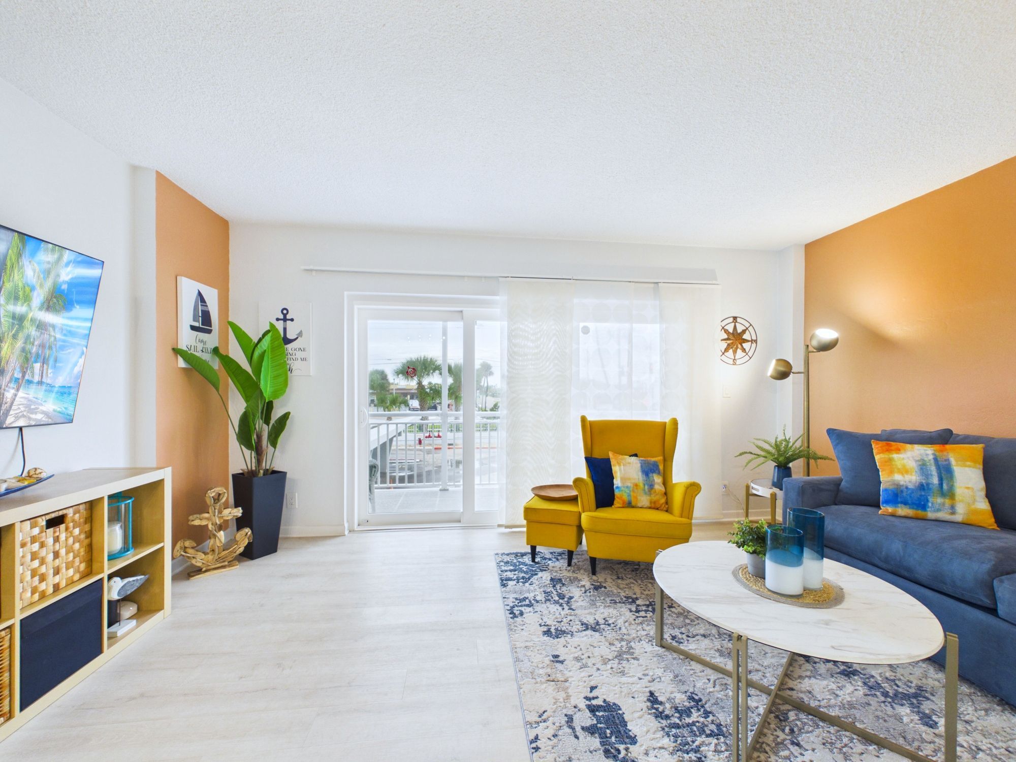 A living room with a yellow armchair, a blue sofa, a white coffee table, and a large window with a view of palm trees.