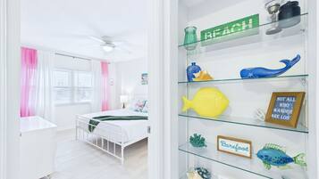 A bedroom with a bed, a white dresser, a window with pink curtains, and a glass shelf with decorative items.