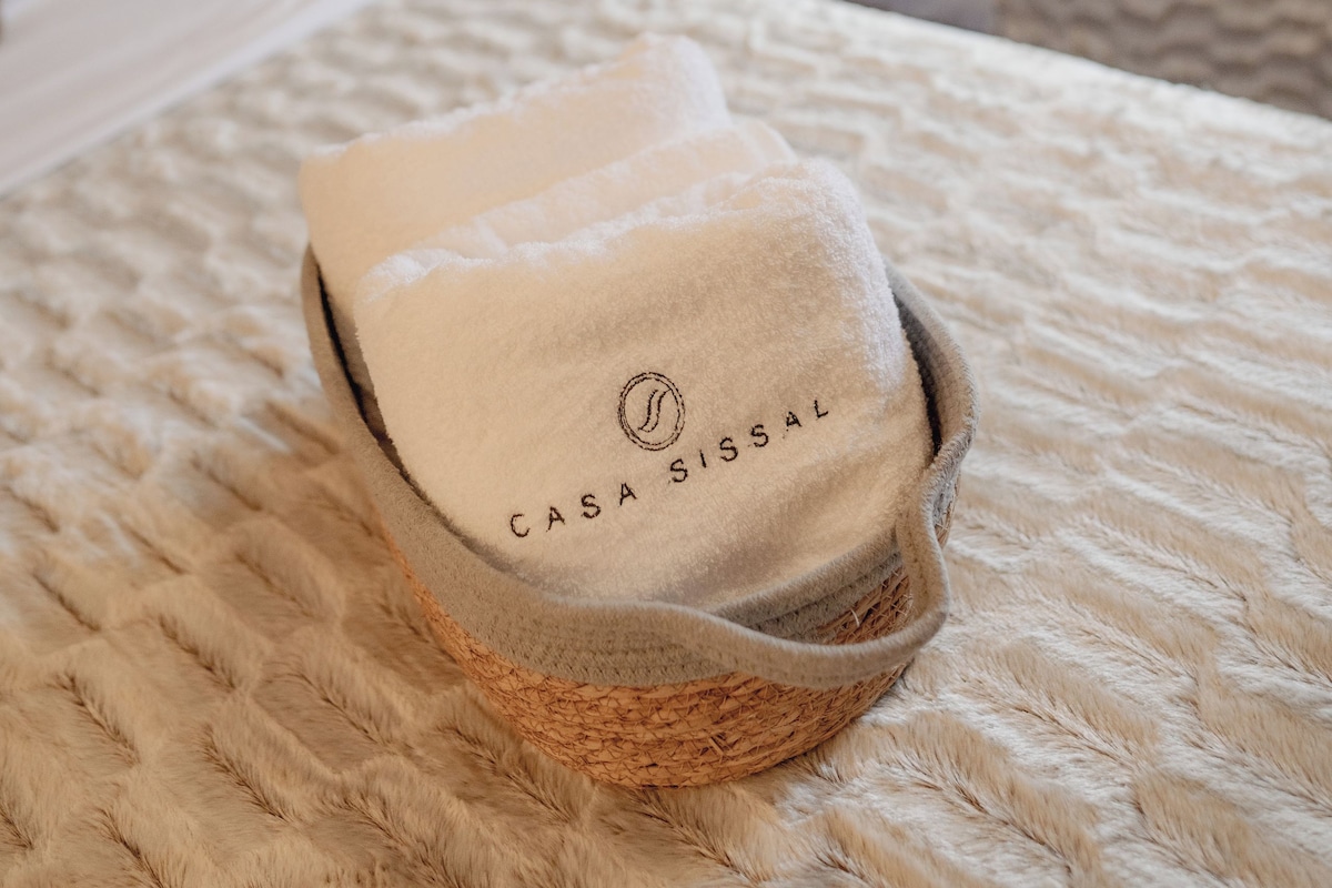 En säng med en flätad korg som innehåller ett hopvikt föremål märkt med texten ”CASAISSAL”.