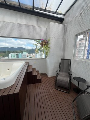A balcony with a wooden floor, a hot tub, a lounge chair, and a view of buildings and mountains.