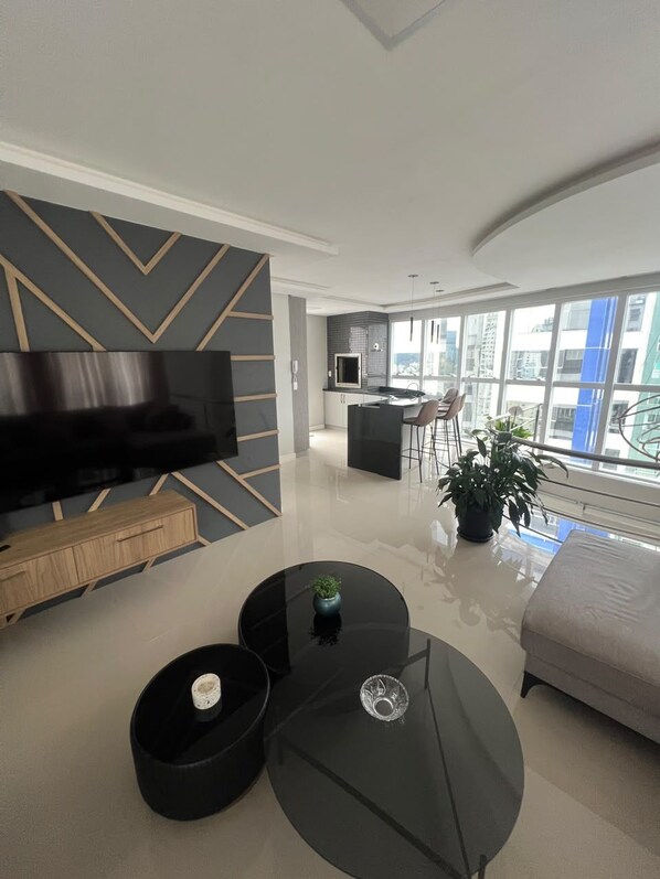 A modern living room with a flat-screen TV, a sofa, a coffee table, and a dining area with chairs.