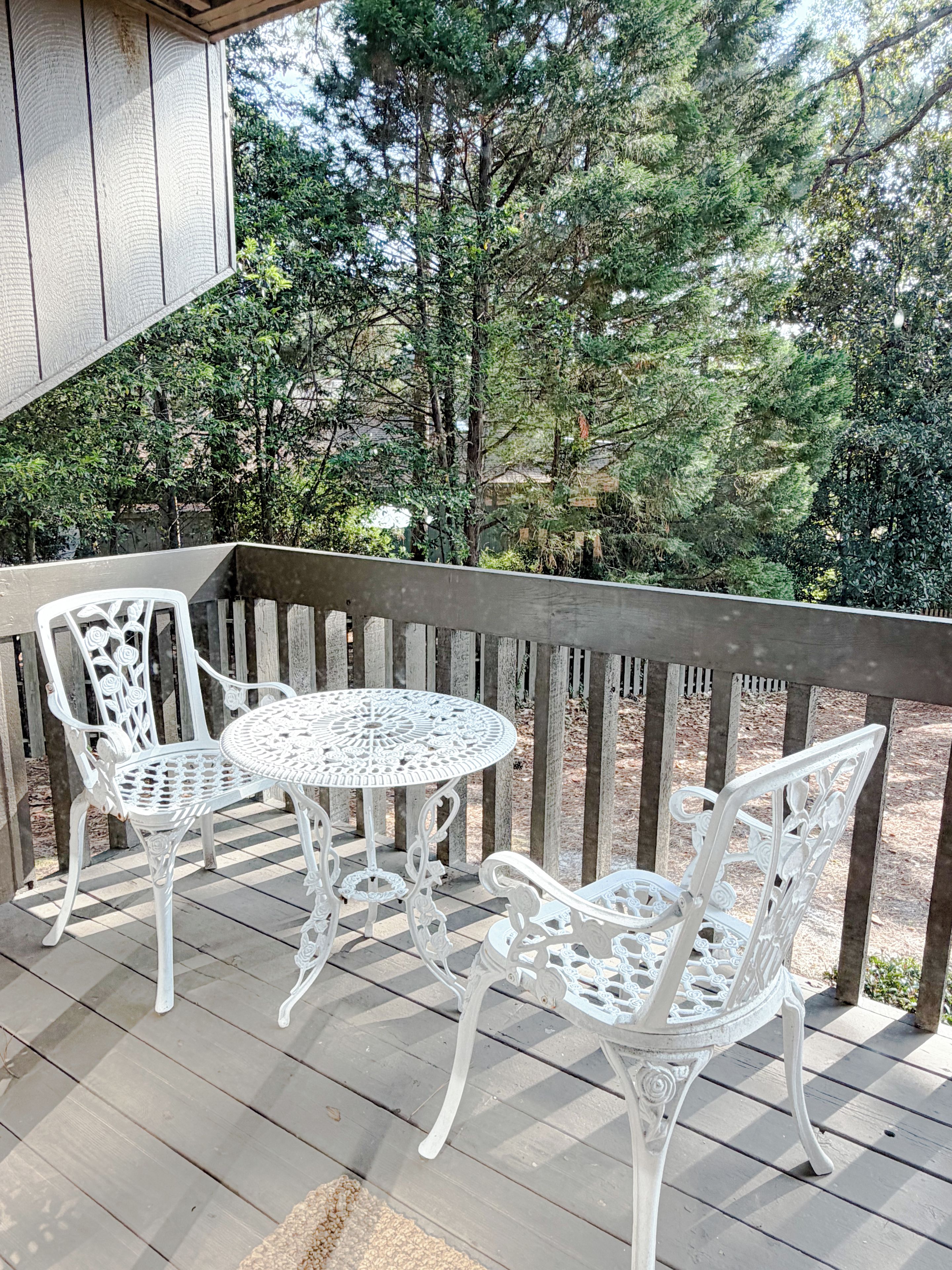 Una terraza de madera con una mesa y sillas de metal blanco, rodeada de árboles.