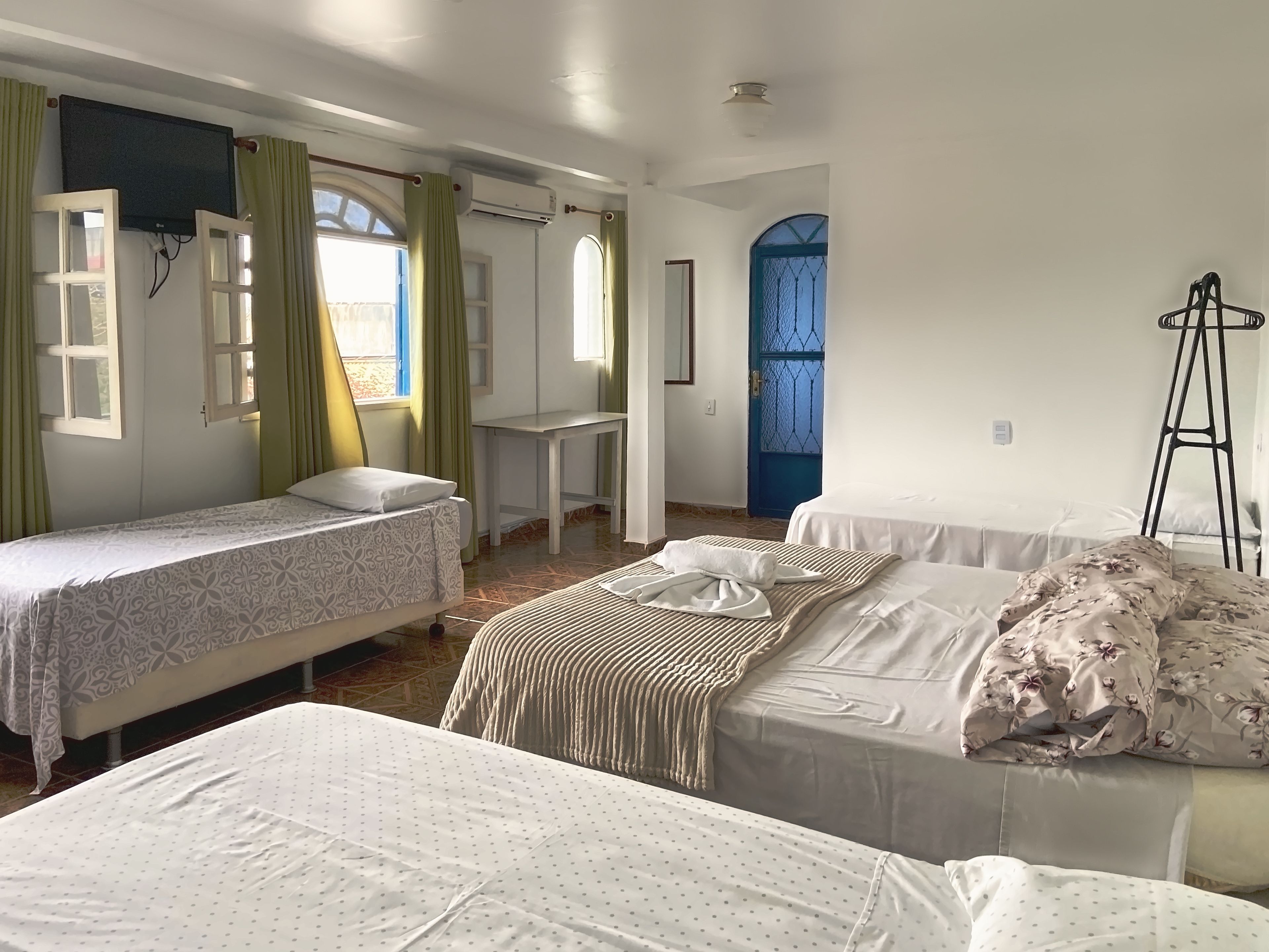 A room with two single beds, a desk, and a chair. There is a window with curtains, a door with a blue gate, and an air conditioning unit on the wall.