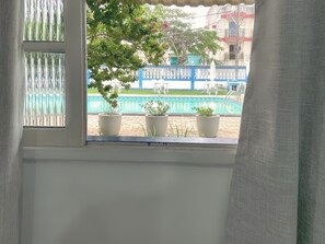 A window with a view of a swimming pool, potted plants, and a building with a blue and white color scheme.