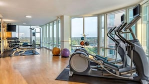 A gym with exercise equipment, a rowing machine, and a view of a cityscape through large windows.