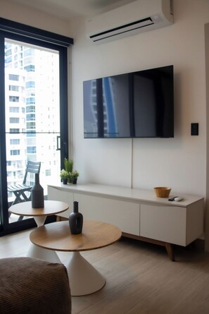 A modern living room with a flat-screen TV mounted on the wall, a round wooden table, a sofa, and a small balcony with a view of buildings. - Modern 1BR with Balcony and Sofa Bed -4 guests (Panamá)