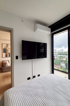 Une chambre d’hôtel moderne dotée d’un téléviseur à écran plat fixé au mur, d’un grand lit recouvert d’une couette blanche et offrant une vue sur le paysage urbain par une grande fenêtre.
