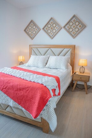 Ein Bett mit hölzernem Gestell, weißer Bettwäsche und einer roten Decke. Zwei Nachttische mit Lampen und einer Wandleuchte über dem Bett.